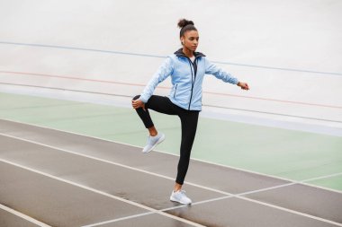 Kablosuz kulaklık takan ve stadyumda esneme egzersizleri yapan güzel ve formda Afrikalı bir sporcu.