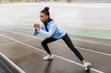 Kablosuz kulaklık takan ve stadyumda esneme egzersizleri yapan güzel ve formda Afrikalı bir sporcu.