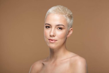 Kısa saçlı, güzel, sarışın bir kadının resmi. Koyu bej duvar arkasında izole edilmiş poz veriyor..