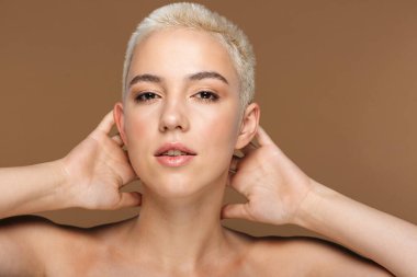 Kısa saçlı, güzel, sarışın bir kadının resmi. Koyu bej duvar arkasında izole edilmiş poz veriyor..