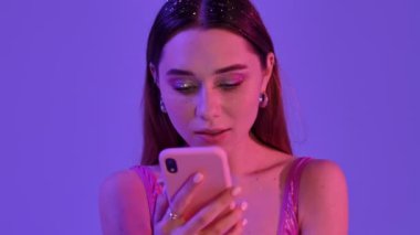 Moda makyajlı mutlu bir kadın menekşe neon arka planında izole edilmiş telefonundan inanılmaz bir haber alırken seviniyor.