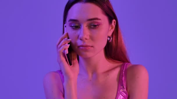 バイオレットネオンの背景に隔離された電話で話している間 ファッションの輝きのメイクを持つ良い表情の感情的な女性が主張しています — ストック動画