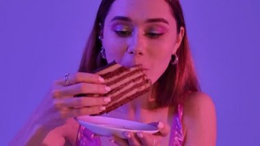 Moda makyajlı mutlu bir kadın lezzetli bir çikolatalı pasta yerken menekşe neon arka planında onu izole ediyor.