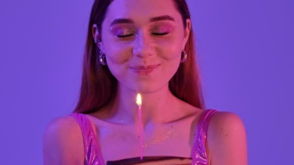 一个快乐快乐的女人 有着时尚艳丽的妆容 她一边在蛋糕里吹着蜡烛 一边唤醒着一个与紫色霓虹灯背景隔离的愿望 — 图库视频影像