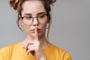 Gri arka planda izole edilmiş gözlük takan genç ve şirin bir kızın yakınına. Sessiz bir jest yapıyor, başka tarafa bakıyor.