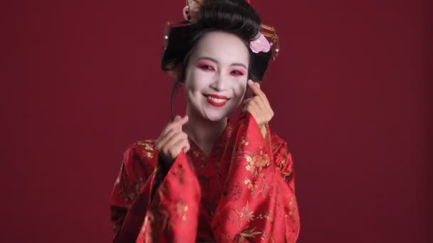 日本の伝統的な着物を着た素敵な笑顔の若い芸者の女性は 赤い背景の上に孤立したジェスチャーで彼女の指で心を示しています — ストック動画