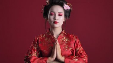 Geleneksel Japon kimonosu giymiş sakin genç bir geyşa kırmızı arka planda izole edilmiş dua pozu gibi el ele tutuşuyor.