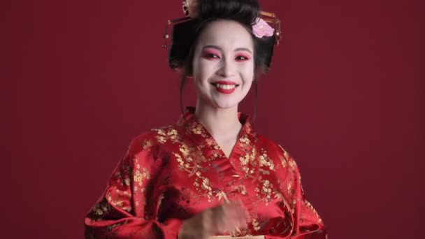 一位穿着日本传统和服 面带微笑的年轻艺妓 在红色的背景下挥动着她的手 — 图库视频影像