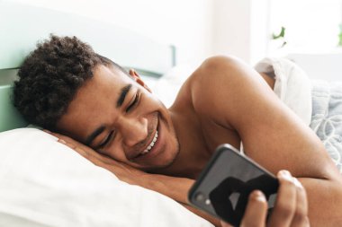 Sabah yatak odasında cep telefonu kullanan neşeli, mutlu, Afrikalı bir genç resmi..