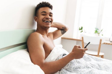 Sabah yatak odasında cep telefonu kullanan neşeli, mutlu, Afrikalı bir genç resmi..