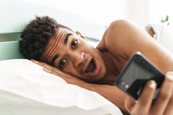 Imagen Sorprendido Joven Africano Sorprendido Mañana Dormitorio Usando Teléfono Móvil —  Fotos de Stock