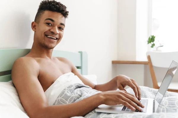 写真の若いです幸せなアフリカ人男でベッドルームで午前中にラップトップコンピュータを使用して — ストック写真