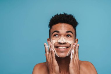 Mavi arka planda izole edilmiş yüz temizleme köpüğü sürerken gülümseyen Afro-Amerikan adamın fotoğrafı.