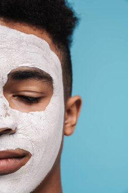 Yüzü kozmetik maskeli sakin Afro-Amerikan bir adamın mavi arka planda gözlerden uzak pozu.