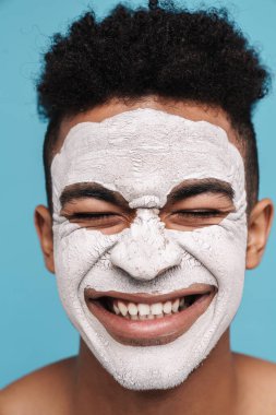 Mavi arka planda izole edilmiş kameraya gülümseyen yüzü estetik maskeli heyecanlı Afrikalı Amerikalı adamın fotoğrafı.