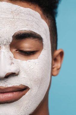 Yüzü kozmetik maskeli sakin Afro-Amerikan bir adamın mavi arka planda gözlerden uzak pozu.