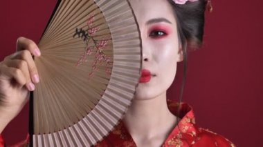 Geleneksel Japon kimonosu giymiş neşeli genç bir geyşa kadının yakın plan görüntüsü kırmızı arka planda izole edilmiş ahşap el yelpazesiyle poz veriyor.