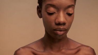 Gizemli, yarı çıplak, kısa siyah saçlı ve bej arka planda kameraya bakan Afrikalı bir kadın.