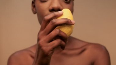 Gülümseyen, yarı çıplak, kısa siyah saçlı, limonla poz veren ve bej arka planda kameraya bakan Afrikalı kadının yakın görüntüsü.