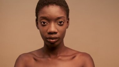 Mutlu, yarı çıplak, kısa siyah saçlı, poz veren ve bej arka planda kameraya bakan Afrikalı kadını yakın çekimde görüyoruz.