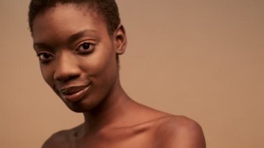 Gülümseyen, yarı çıplak, kısa siyah saçlı, bej arkaplanına bakıp duran Afrikalı bir kadın.