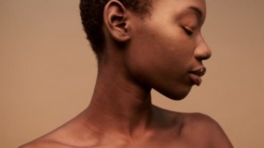 Gizemli, yarı çıplak, kısa siyah saçlı Afrikalı bir kadın, etrafa bakıyor ve bej arka planda sessizlik işareti yapıyor.