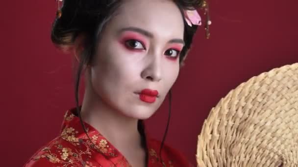 一个穿着日本传统和服的漂亮的年轻艺妓女子的特写镜头 是用木扇把它从一边移动到另一边 在红色的背景下与另一边隔开 — 图库视频影像