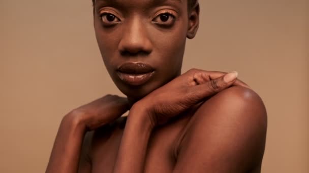 Close up view of Sensual pretty half-naked african woman with short black hair holding hands on her shoulders and looking around over beige background