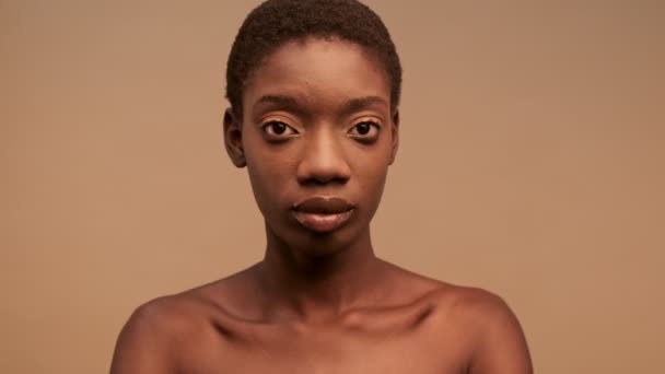 アップビューの静かなかなり半分裸アフリカ人女性とともに短い黒髪ポーズとカメラを見て上のベージュの背景 — ストック動画