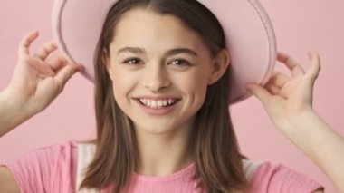 Mutlu esmer kadın, tulumlu, şapkalı, poz veren ve pembe arka planda kameraya bakan.