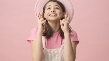 Tulumlu ve şapkalı güzel bir kadın dua ediyor, parmaklarını çapraz yapıp pembe arka plana bakıyor.