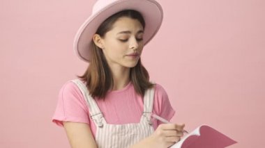 Tulum giymiş, şapkalı güzel bir kadın birini dinliyor ve pembe arka plan üzerine deftere bir şeyler yazıyor.
