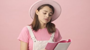 Tulumlu ve şapkalı güzel esmer kadın pembe arka planda deftere bir şeyler okuyor.