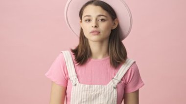 Hoş olmayan esmer kadın tulumlu ve pembe arka planda yüzünü kapatan şapkalı.