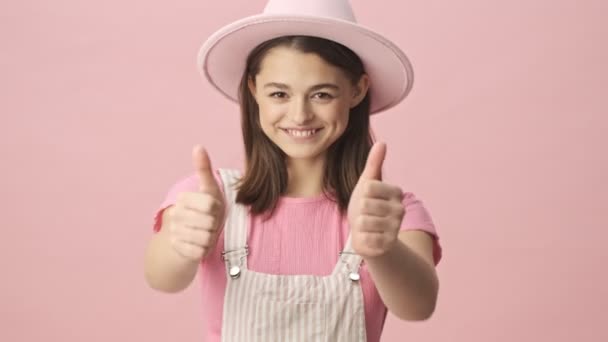 幸せなかわいいですブルネット女性で全体と帽子ショー親指アップと見ますザカメラ上のピンクの背景 — ストック動画