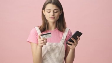 Tulum giymiş, gülümseyen esmer kadın. Pembe arka planda kredi kartı ve akıllı telefonuyla faturaları ödüyor.