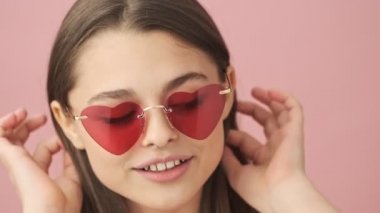 Tulum giymiş, güneş gözlüklü, gülümseyen esmer kadının yakın görüntüsü. Saç stilini düzeltiyor ve pembe arka plandan kameraya bakıyor.