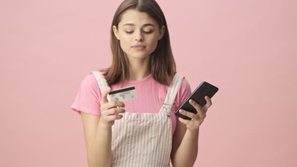 Улыбающаяся Брюнетка Комбинезоне Оплачивает Счета Кредиткой Смартфоном Розовом Фоне — стоковое видео