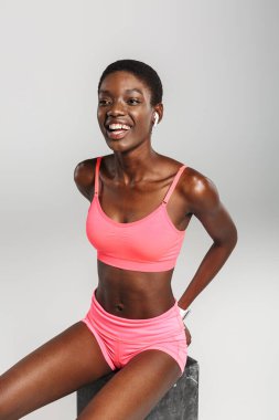 Beyaz arka planda izole bir şekilde otururken kablosuz kulaklık takan gülen Afrikalı Amerikalı spor kadını görüntüsü