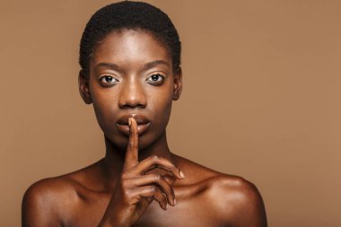 Bej arka planda izole edilmiş dudaklarda parmak tutan çekici, yarı çıplak Afrikalı bir kadının portresi.