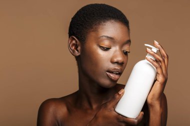 Bej arka planda izole edilmiş vücut losyonu şişesi tutan güzel, yarı çıplak Afrikalı bir kadının portresi.