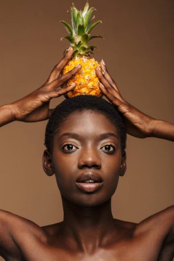 Kafasında ananas tutan güzel, yarı çıplak bir Afrikalı kadının portresi kahverengi arka planda izole edilmiş.