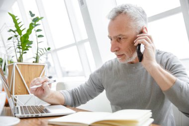 Ciddi, olgun bir adamın cep telefonu ve laptop ile kapalı bir kafede çalışırken not aldığı bir fotoğraf.
