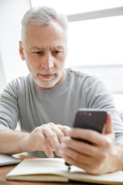 Ciddi, olgun bir adamın, kafeteryada otururken cep telefonuyla yazı yazıp not aldığı fotoğraf.