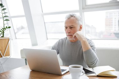 Odaklanmış yetişkin bir adamın kahve içerken ve laptopla çalışırken çekilmiş fotoğrafı.