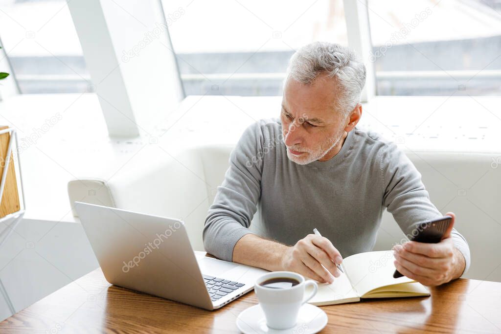 Photo of focused mature man making notes while working with laptop and cellphone in cafe indoors