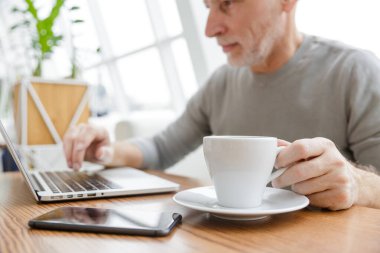 Odaklanmış yetişkin bir adamın kahve içerken ve laptopla çalışırken çekilmiş fotoğrafı.