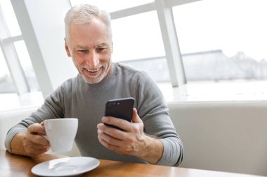 Kahve içen ve kafede otururken akıllı telefon kullanan neşeli, olgun bir adamın fotoğrafı.