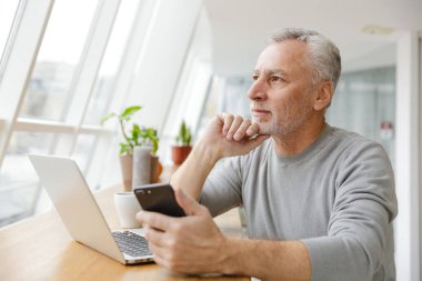 Ciddi bir yetişkin adamın kahve içerken cep telefonu ve laptopuyla kapalı bir kafede çalışırken çekilmiş fotoğrafı.