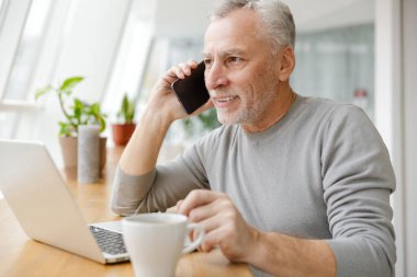 Gülümseyen olgun bir iş adamının görüntüsü kafede oturmuş cep telefonuyla dizüstü bilgisayar kullanarak konuşuyor..
